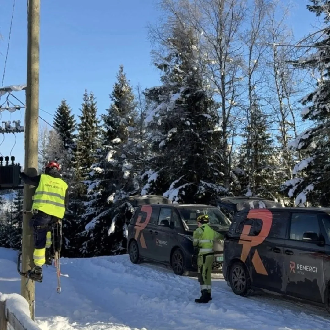 Person i opplæring oppe i en stolpe en vinterdag, med to av Renergis biler i snøen under