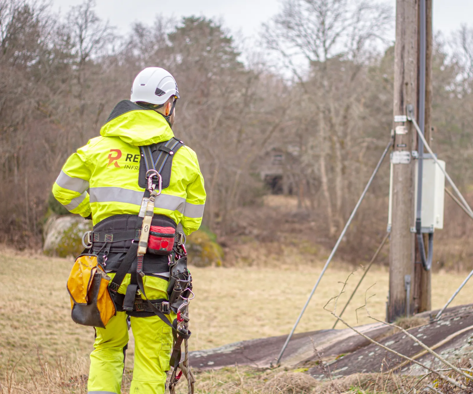 Montør fra Renergi Infra med klatresele og verktøy går over en mark mot en trestolpe med et skap